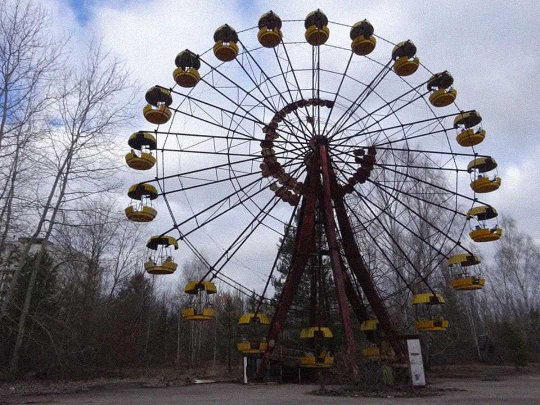 An abandoned Ferris wheel with yellow passenger cabins stands in an overgrown, deserted area surrounded by leafless trees under a cloudy sky. The scene looks neglected and eerie.