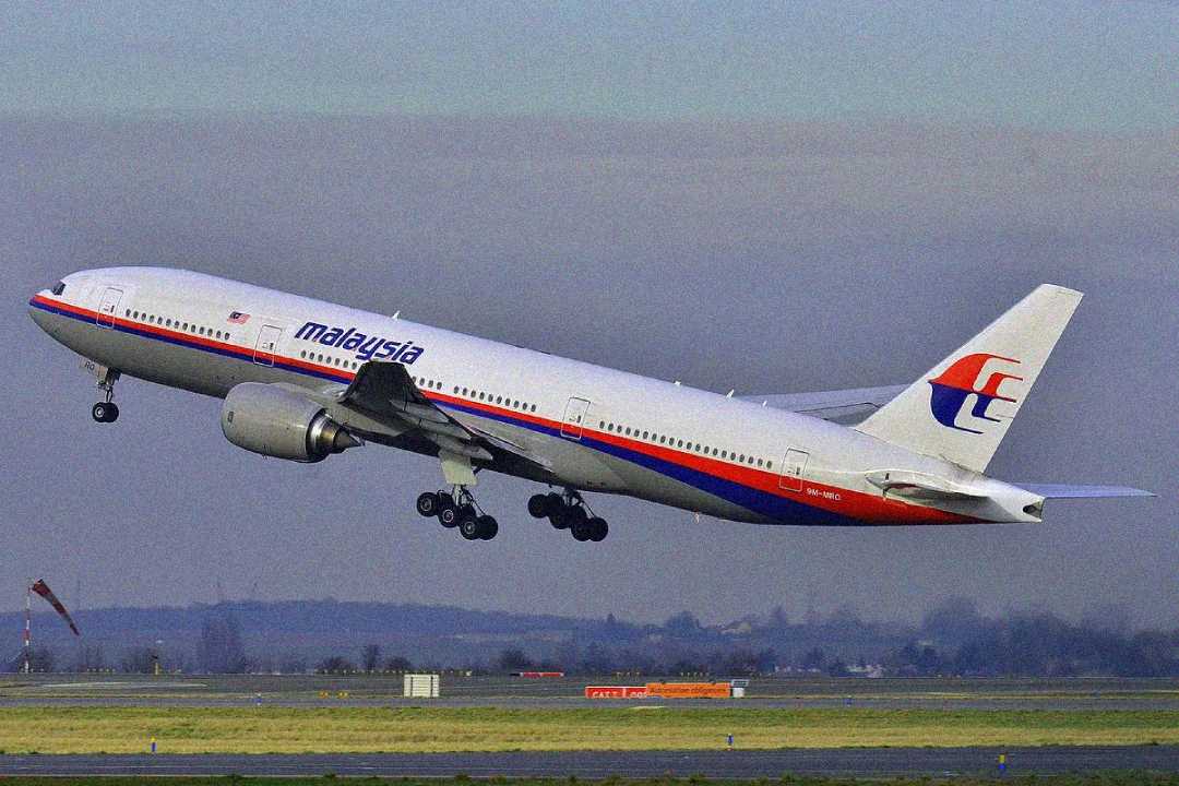 A Malaysia Airlines passenger jet takes off from a runway, with its landing gear still extended. The aircraft is white with red and blue stripes and the Malaysia Airlines logo on the tail. The sky is overcast.