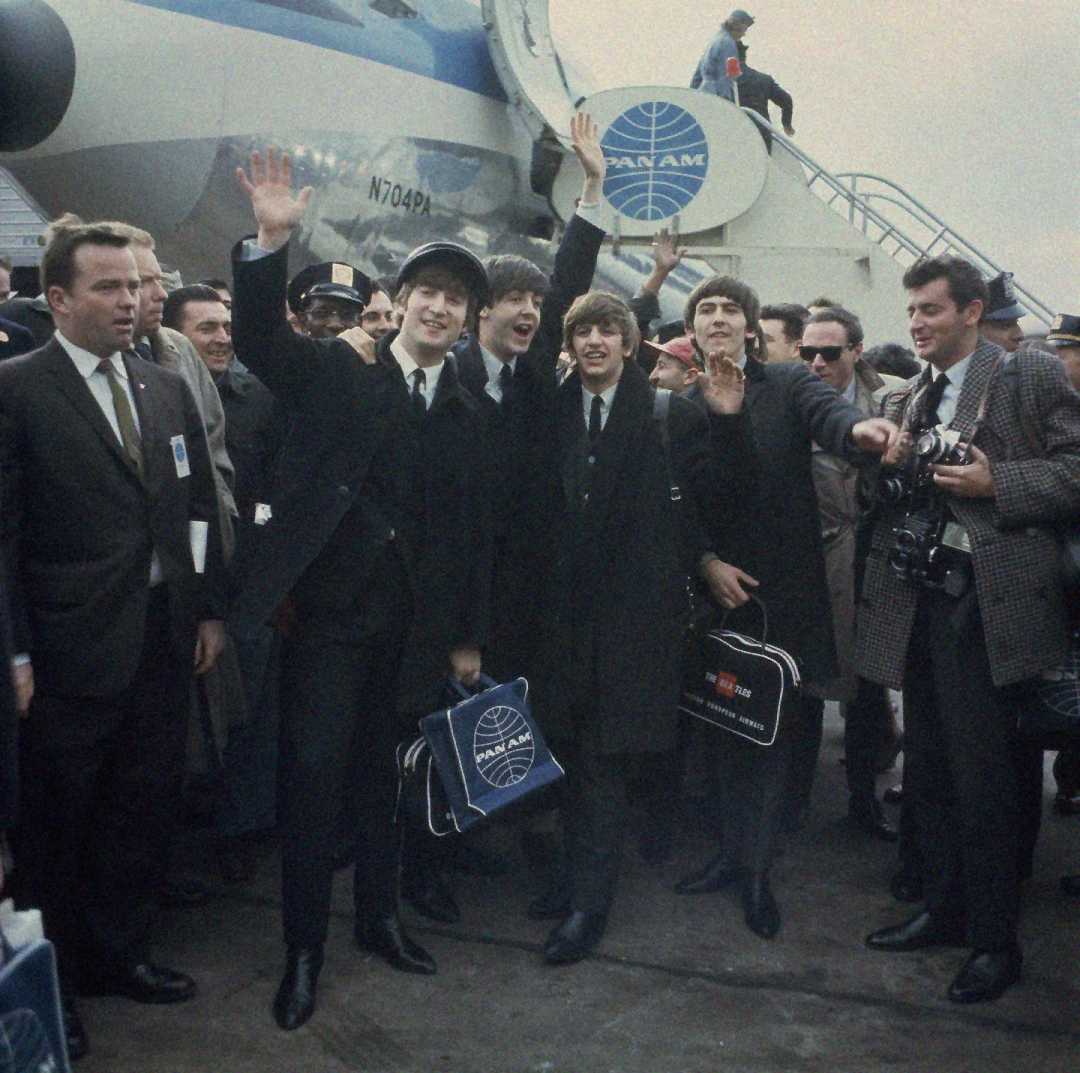 The Beatles, dressed in suits, wave and smile as they arrive at an airport, carrying Pan Am bags, surrounded by officials, photographers, and fans near a Pan Am airplane.