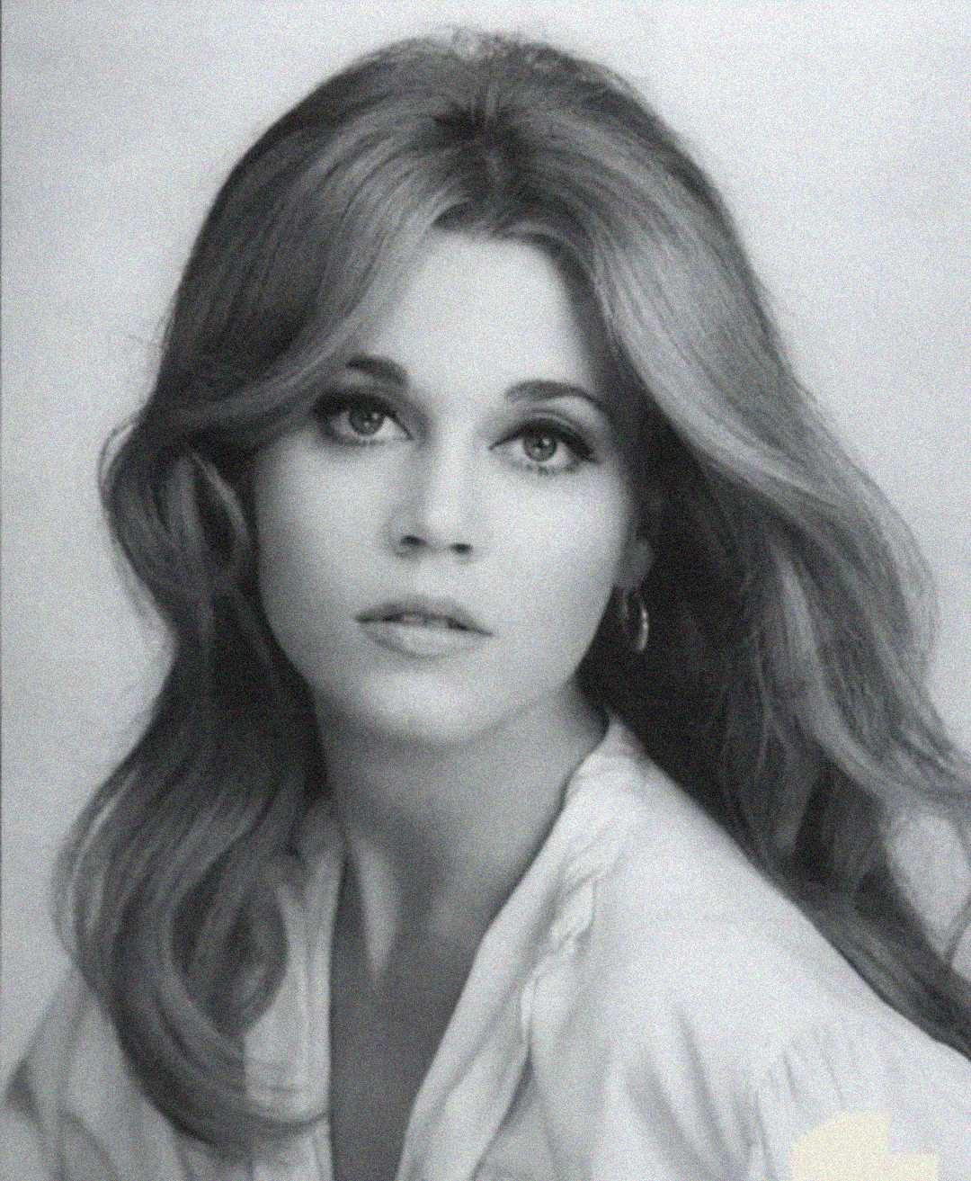 Black and white portrait of a young woman with long wavy hair, wearing a light-colored top and hoop earrings, looking directly at the camera with a calm expression.