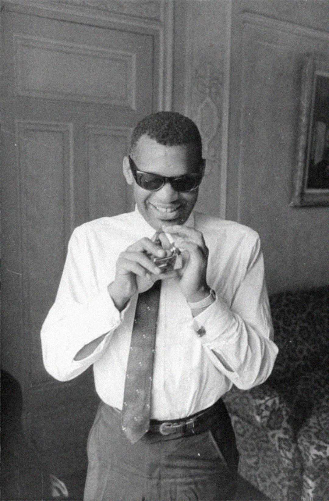 A man wearing sunglasses and a tie smiles while holding a small glass object with both hands in a vintage-style room with patterned wallpaper and a framed picture on the wall.