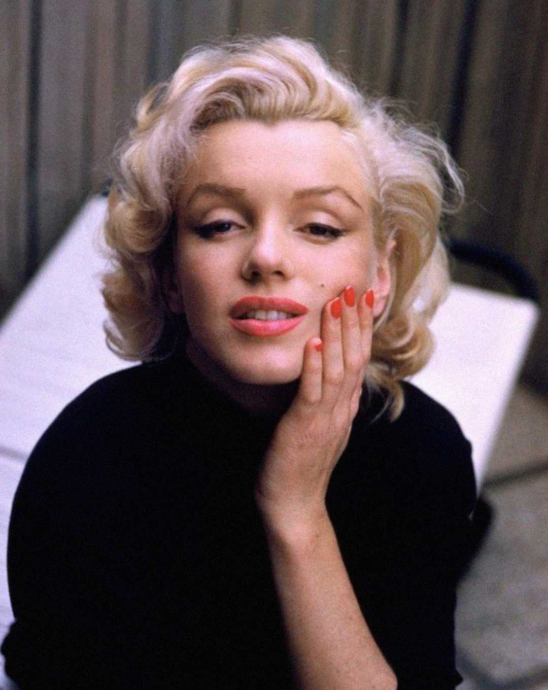 A woman with short, wavy blonde hair and red lipstick rests her hand on her cheek, wearing a black top and red nail polish, looking slightly up with a gentle expression. A blurred background is visible.