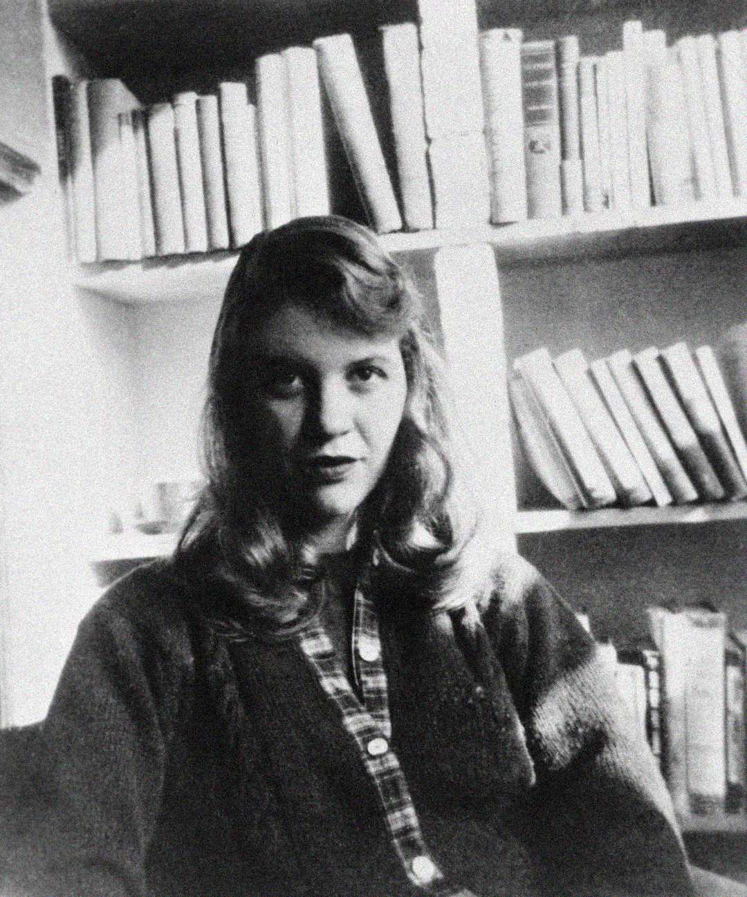 A black-and-white photo of a young woman with long hair, wearing a plaid shirt and cardigan, sitting in front of bookshelves filled with books. She looks directly at the camera.