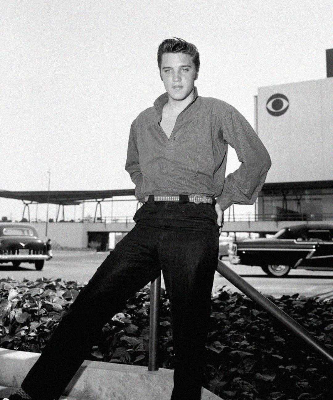A young man with dark hair, wearing a button-down shirt and dark pants, stands confidently outdoors near classic cars and a building with a CBS logo, in a black-and-white photo.