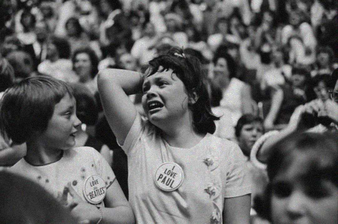 A black-and-white photo of two young girls in a crowd, one smiling and the other appearing emotional. Both wear T-shirts with badges—one reads "Love The Beatles" and the other "Love Paul." The background is filled with people.