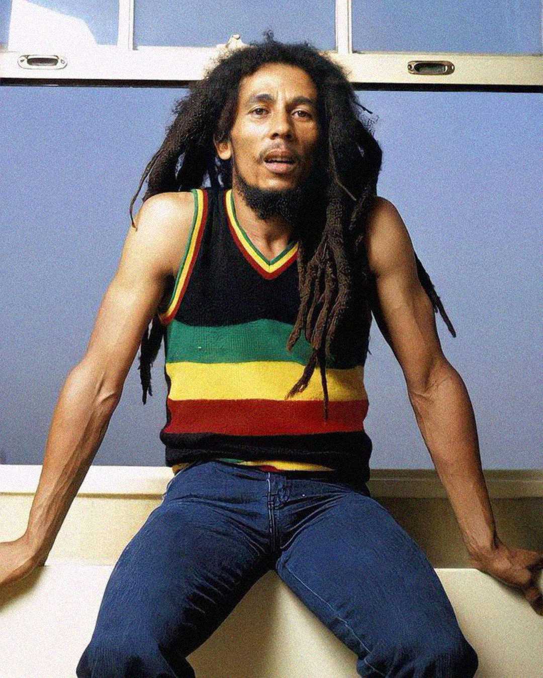 A man with long dreadlocks sits on a ledge in front of a window, wearing a sleeveless black shirt with red, yellow, and green stripes, and blue jeans.
