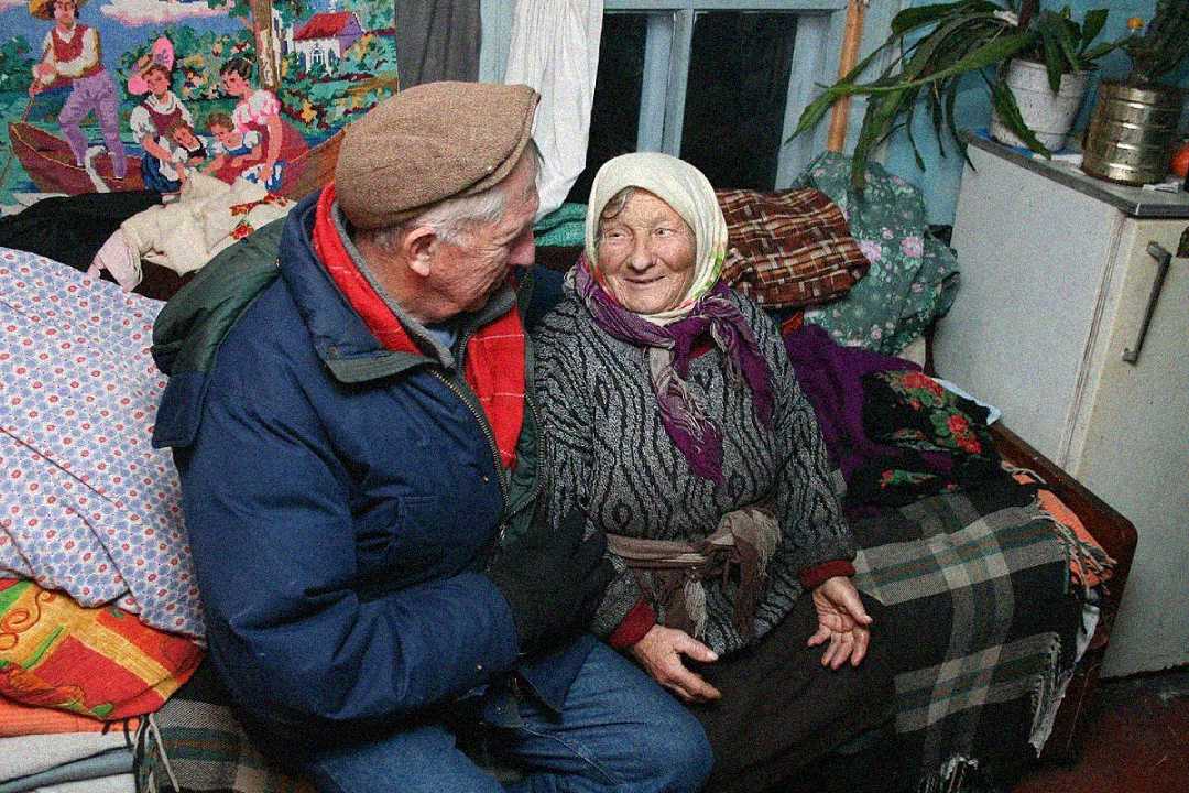 An elderly man and woman sit together on a colorful, cozy bed in a warmly decorated room, smiling and holding hands. Blankets, a painting, and plants add to the homey atmosphere.