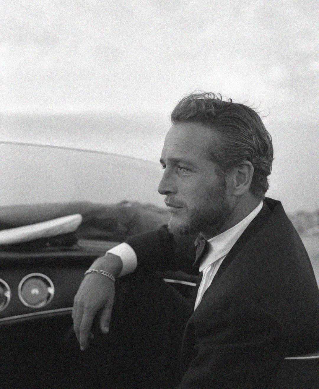 A black-and-white photo of a man in a suit sitting in a vintage car, looking thoughtfully into the distance. His hair is slicked back and he has a short beard. The background is blurred, highlighting the subject.