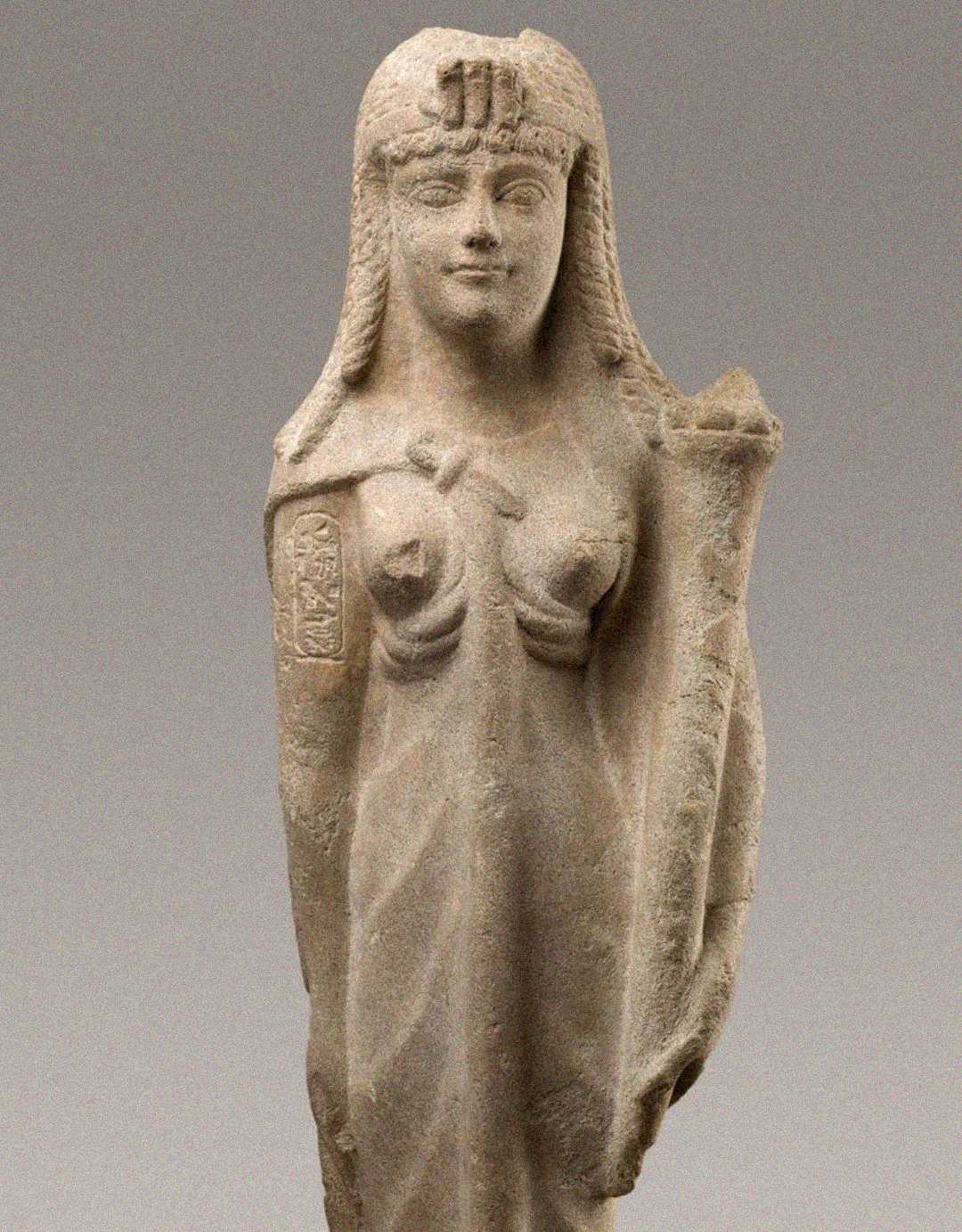 Ancient stone statue of a woman with long headdress, wearing a draped garment. Her left arm is bent and her right arm hangs by her side. The surface shows some wear and missing features.