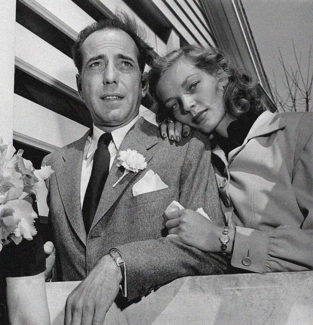 A man in a suit with a flower in his lapel stands next to a woman in a coat, who leans on his shoulder. They are outdoors, close to a building, looking slightly away from the camera.