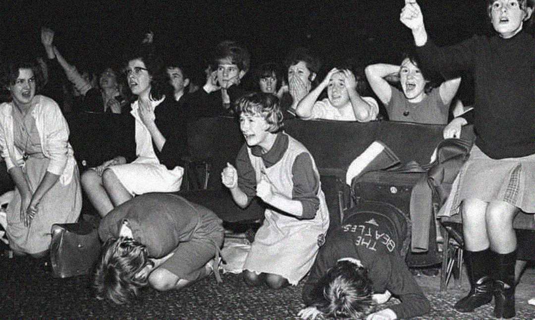 Black-and-white photo of a group of young women in an audience, some standing, some sitting, and others crouched on the floor, all appearing extremely excited and emotional, possibly screaming or covering their faces.