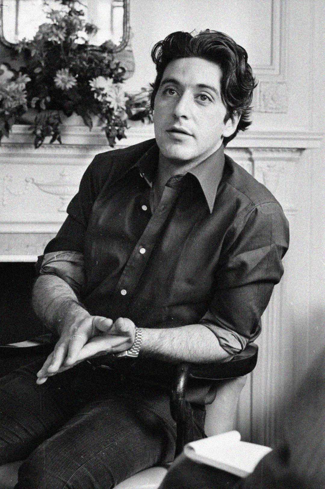 A man with dark, wavy hair sits in a chair wearing a dark button-up shirt and watch, gesturing with his hands. Behind him is a fireplace with a flower arrangement. The image is in black and white.