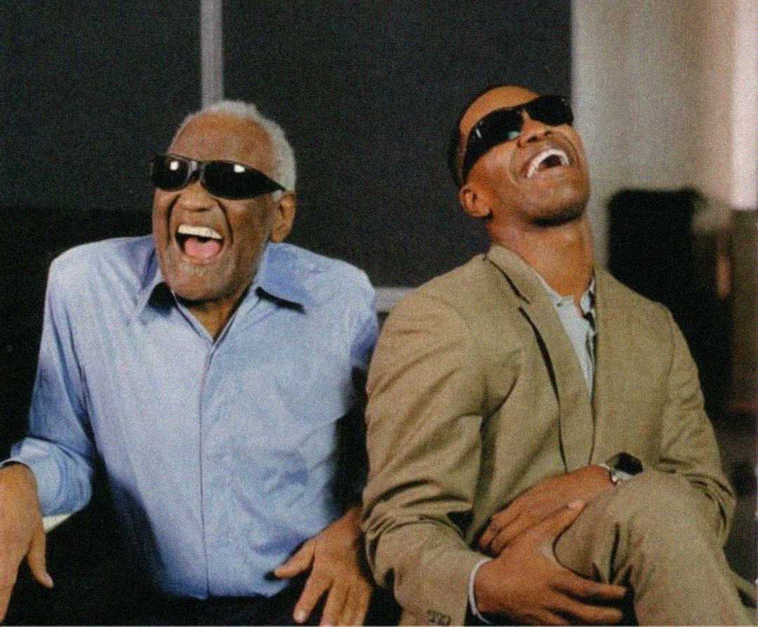 Two men wearing sunglasses laugh together while sitting side by side. One is older with gray hair and a blue shirt, the other is younger in a tan suit. Both appear joyful and relaxed in a bright indoor setting.