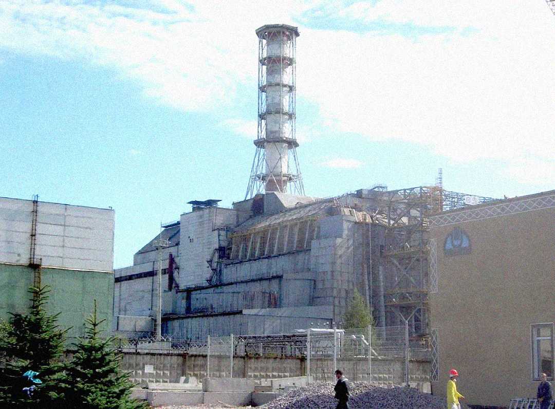 The image shows the damaged Chernobyl nuclear power plant reactor building, with scaffolding and containment structures. Two people in protective clothing stand in the foreground near a fence and small trees under a bright sky.