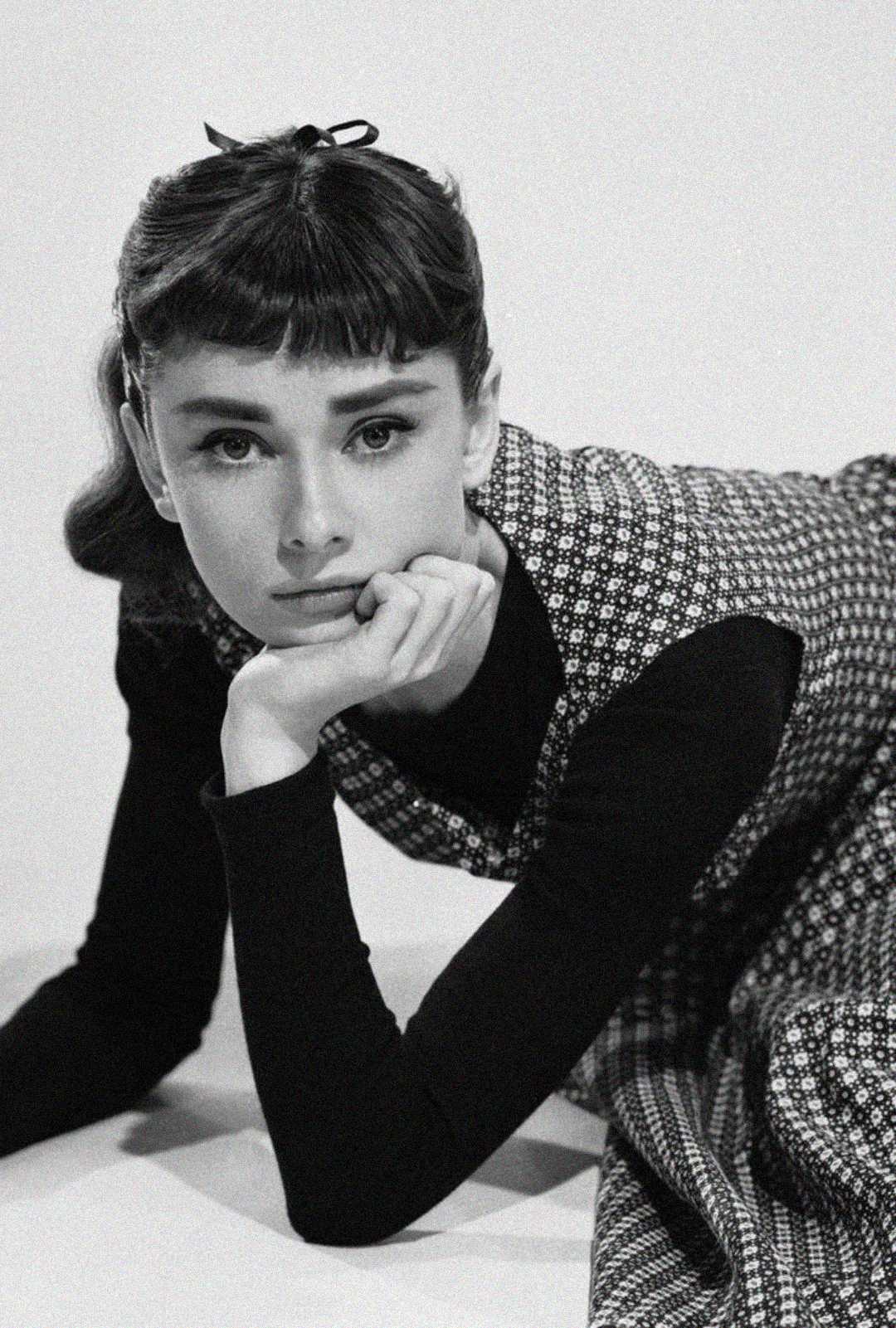 Black and white photo of a young woman with short, dark hair and bangs, wearing a patterned dress over a long-sleeve shirt, resting her chin on her hand and looking thoughtfully at the camera.
