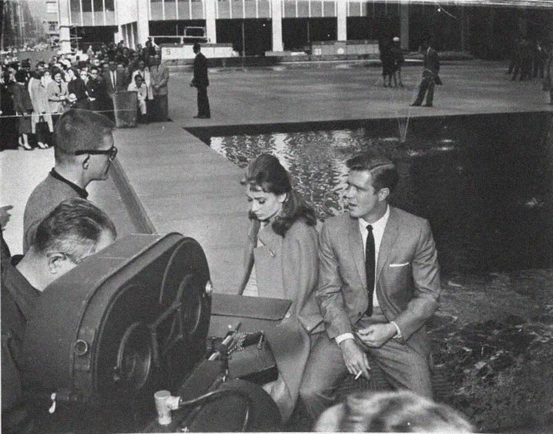 A black-and-white photo shows a woman and a man in formal clothing sitting by a reflective pool, surrounded by film equipment and crew, with a crowd watching in the background.