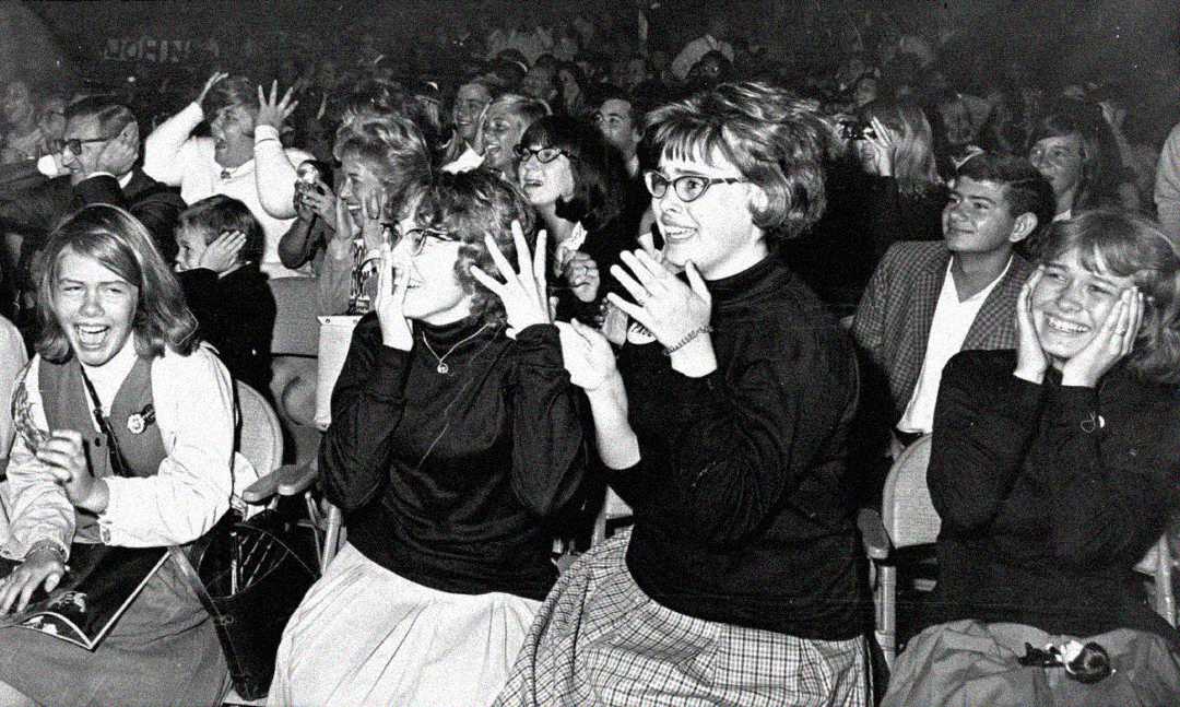 A black and white photo of an excited crowd, mostly young women, smiling, laughing, and holding their faces in awe while sitting in rows at a concert or public event.