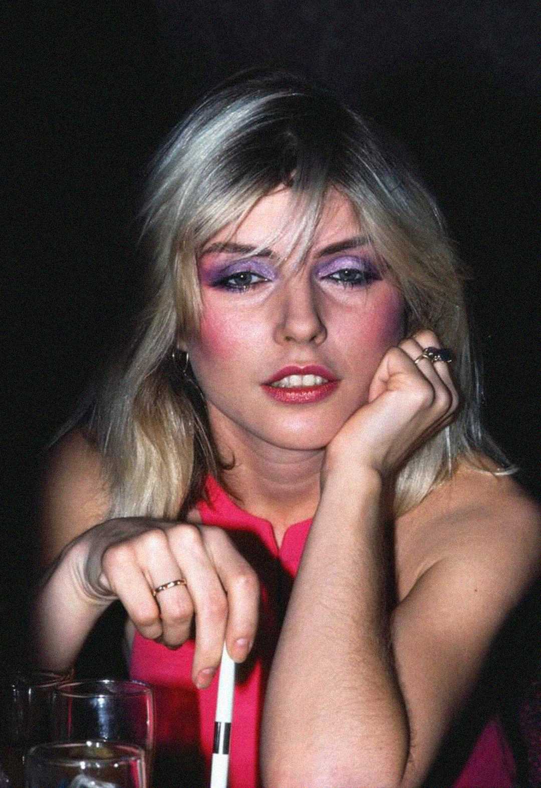 A woman with blonde hair and bold pink and purple makeup rests her chin on her hand while holding a straw. She wears a sleeveless pink top and several rings, sitting at a table with drinks in dim lighting.