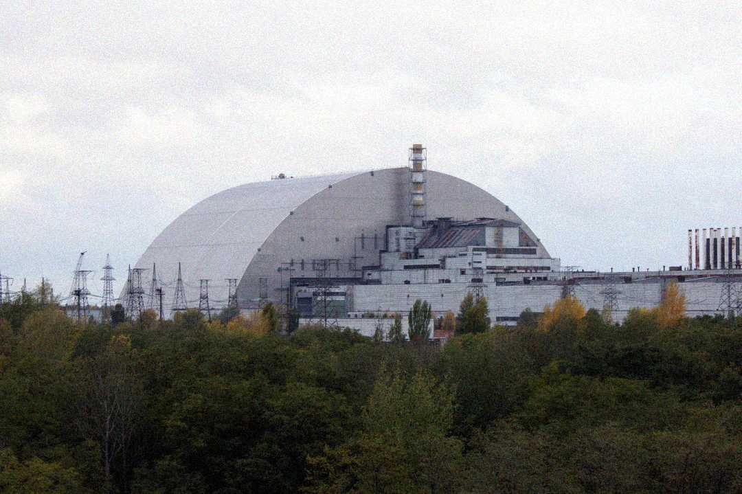 A large, arched structure covers the remains of a damaged industrial building surrounded by trees, under a cloudy sky. Several smokestacks and power lines are visible around the complex.