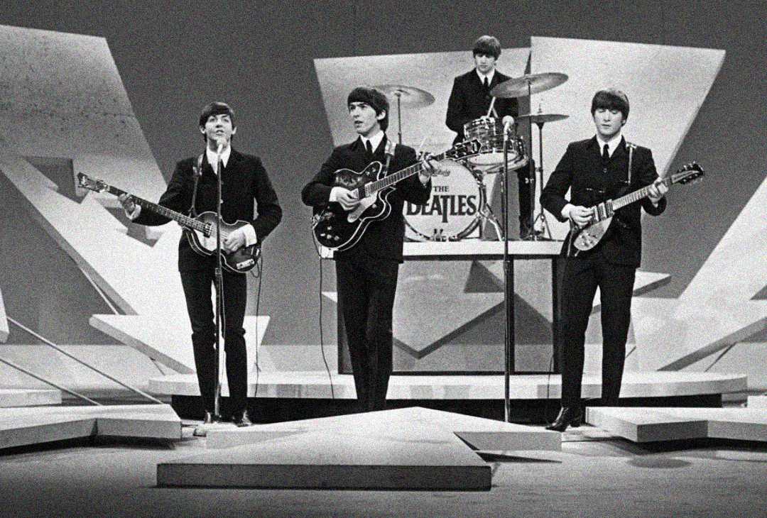 Four musicians in suits perform on a geometric stage. Three play guitars and sing at the front, while a drummer plays behind them. The drum kit displays “The Beatles.” The scene is in black and white.