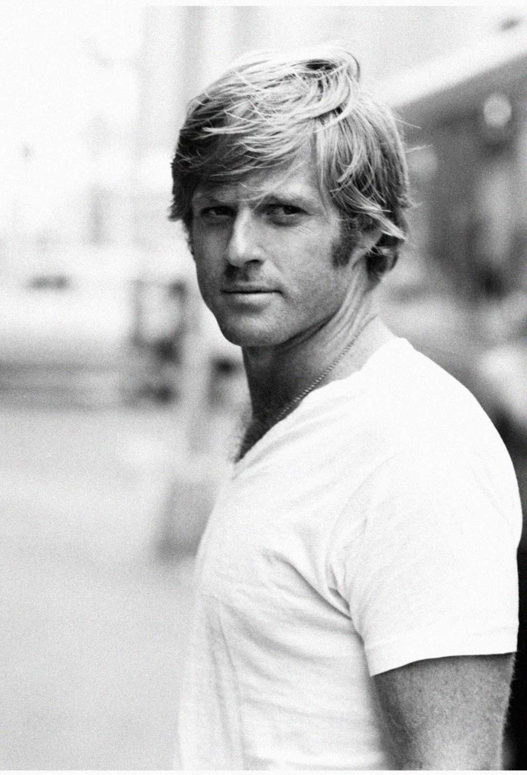 Black and white photo of a man with light hair, wearing a white T-shirt, standing outdoors and looking over his shoulder toward the camera with a neutral expression. The background is blurred.