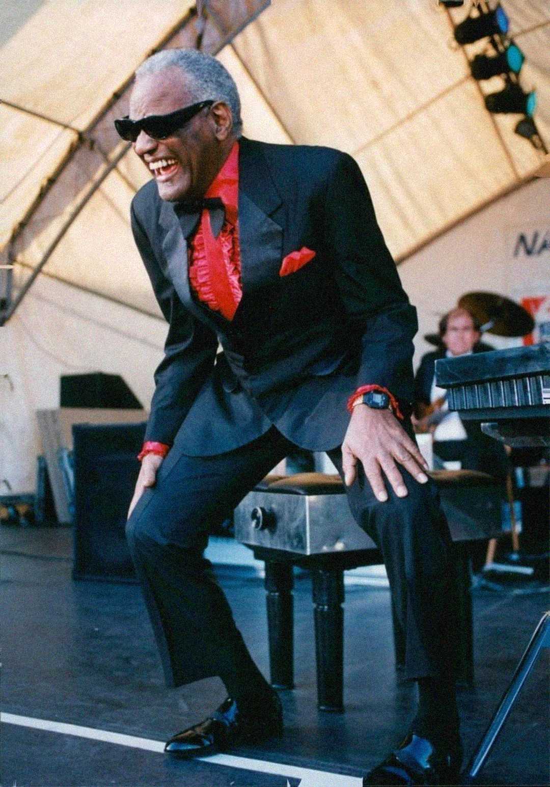 An older man in a black suit with red accents and sunglasses smiles broadly while standing by a piano on an outdoor stage, with musicians and equipment visible in the background.