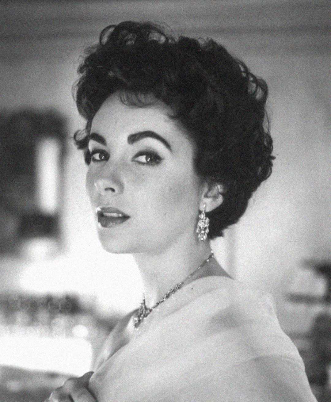 Black and white portrait of a woman with short curly dark hair, wearing earrings and a necklace, looking over her shoulder towards the camera with a poised expression. The background is softly blurred.