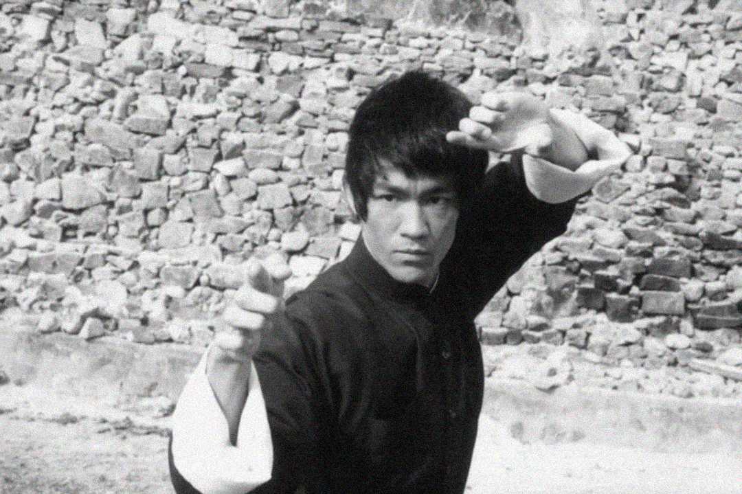 A person in martial arts attire strikes a defensive pose with hands raised in front of a stone wall background, appearing focused and intense.