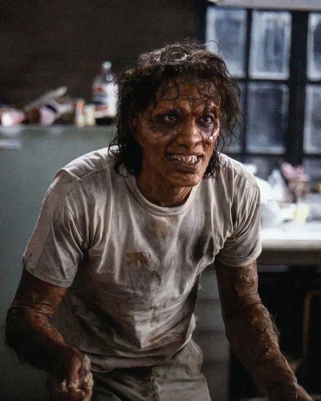 A disheveled person with messy hair, dirty skin, and torn, stained clothes stands in a dimly lit room, appearing distressed and wild-eyed, with a cluttered counter and window in the background.