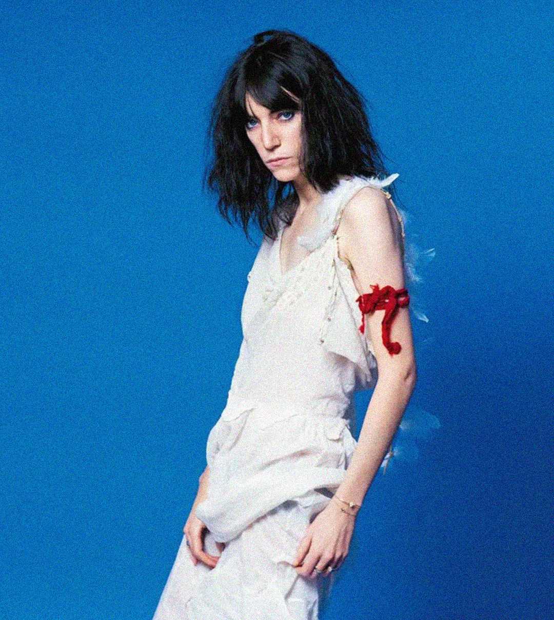 A woman with dark, tousled hair wears a white, ruffled dress and a red ribbon tied around her upper arm, standing against a solid blue background. She has a serious expression and gazes toward the camera.
