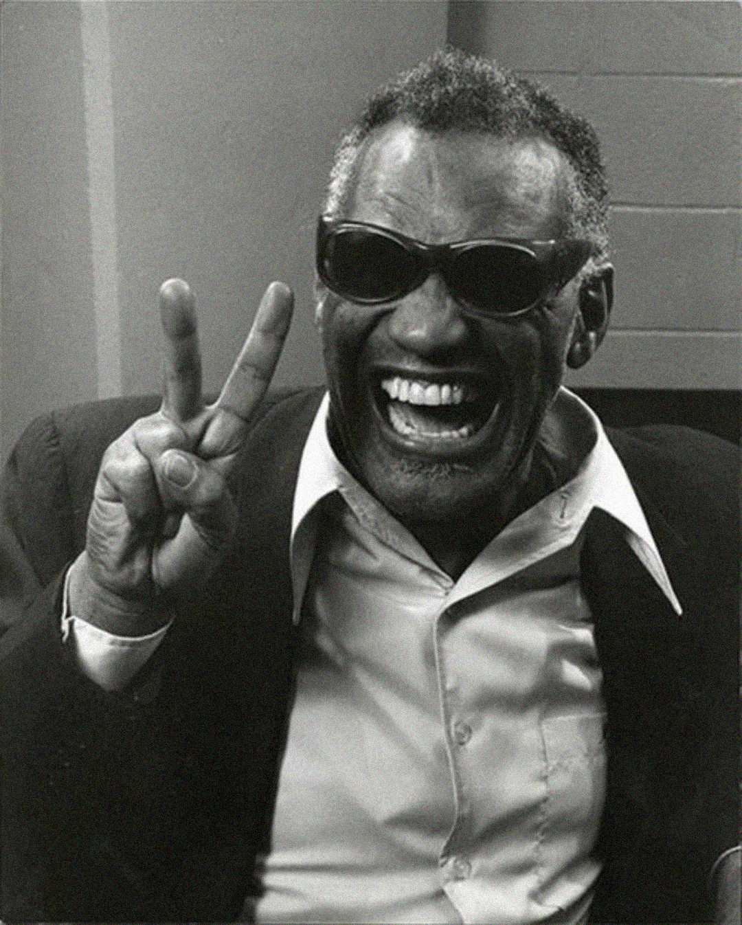 A man wearing dark sunglasses smiles broadly while holding up his fingers in a peace sign. He is dressed in a collared shirt and blazer, sitting indoors. The image is in black and white.