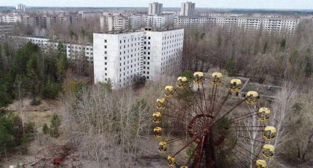 An abandoned Ferris wheel stands amid overgrown trees and decaying buildings in a deserted city, with several high-rise apartments visible in the background.