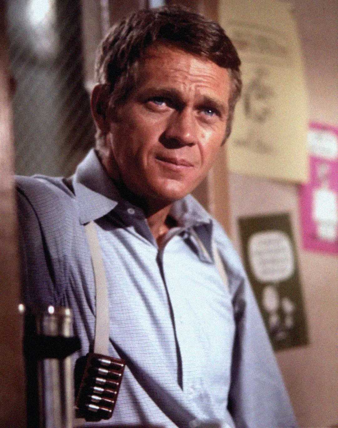 A man with short brown hair wearing a light blue button-up shirt and suspenders stands indoors, looking slightly to the side with a serious expression. There are posters and a window behind him.