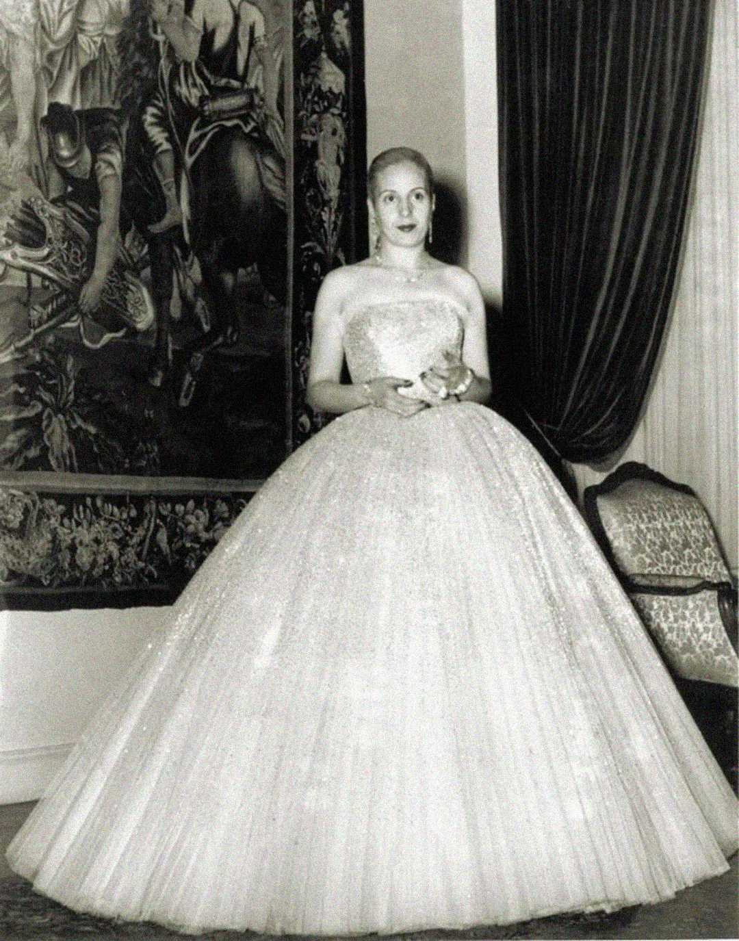 A woman in a strapless, full-skirted ball gown stands indoors near a tapestry and a curtained window, holding a small object and facing the camera in this black and white photograph.