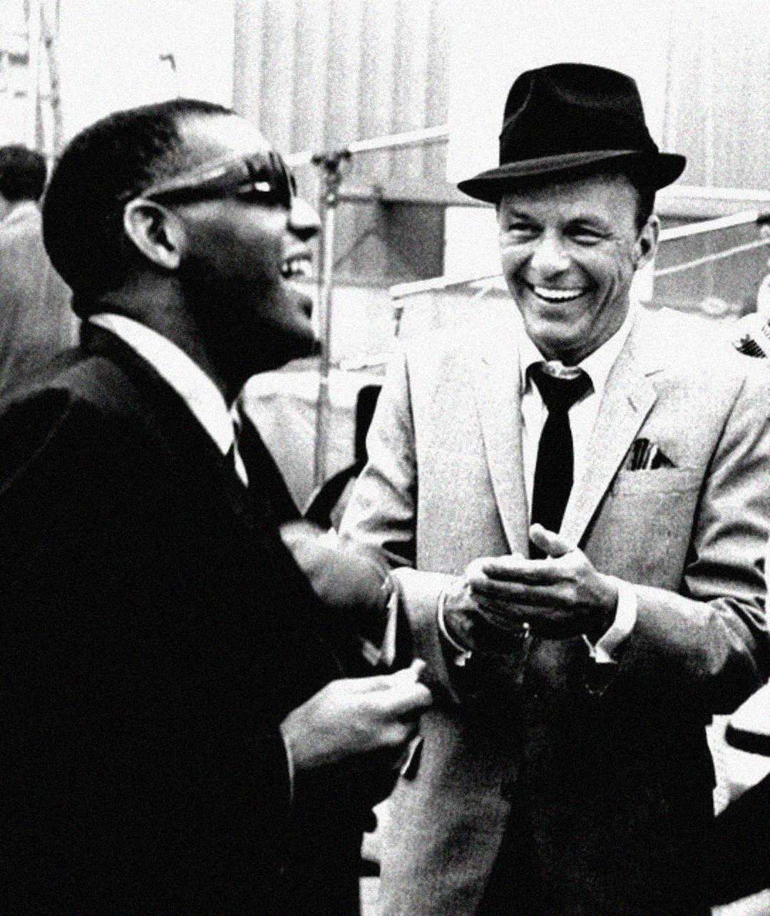 Two men in suits and ties share a laugh. One wears sunglasses, while the other wears a hat and claps his hands. Both appear to be enjoying a lighthearted moment together. The image is black and white.