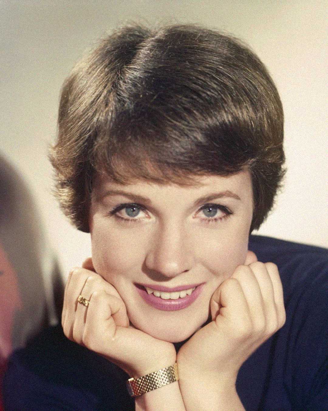 A young woman with short brown hair and blue eyes smiles gently, resting her chin on her hands. She wears a gold wristwatch and a ring, with a soft, light background behind her.