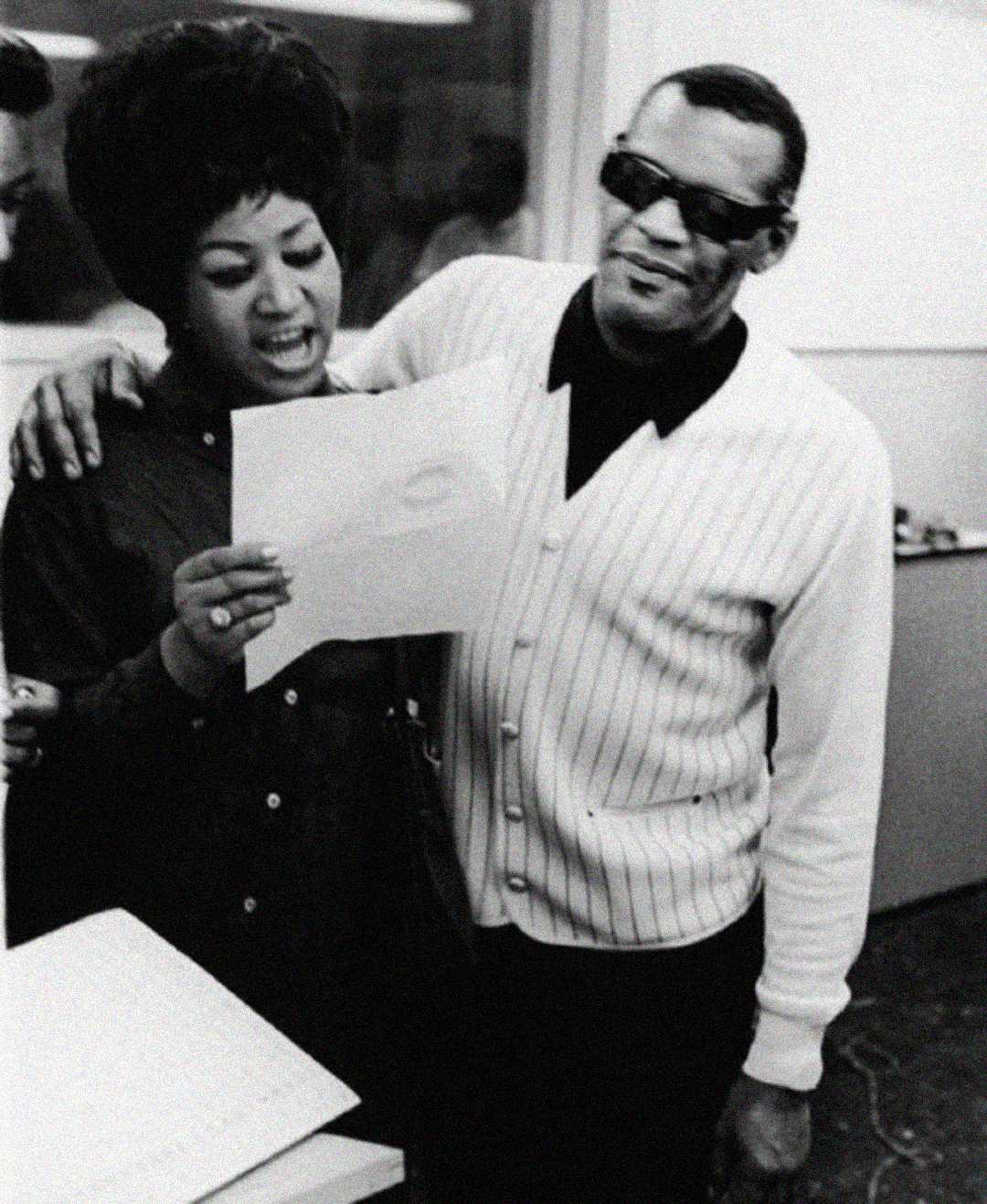 A woman and a man stand together in a recording studio. The woman is holding and reading a sheet of paper, while the man, wearing sunglasses and a striped sweater, smiles with his arm around her shoulders.