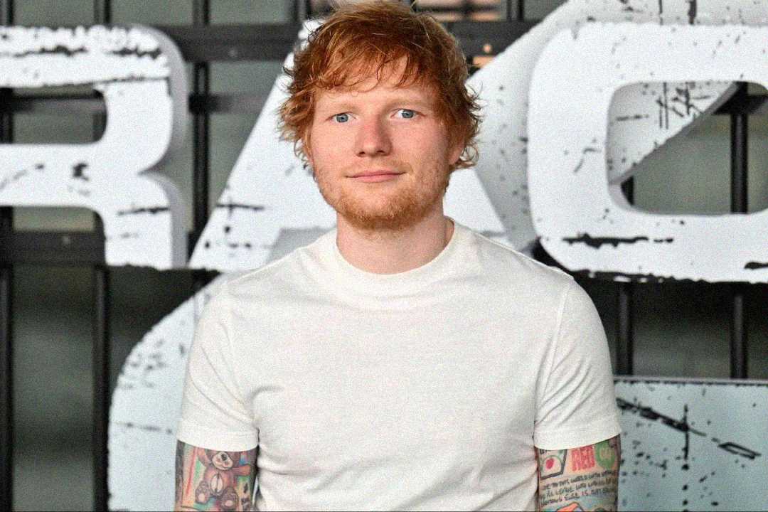 A man with fair skin, red hair, and a short beard stands in front of a large, partially visible white sign. He is wearing a plain white t-shirt and has colorful tattoos on both arms.