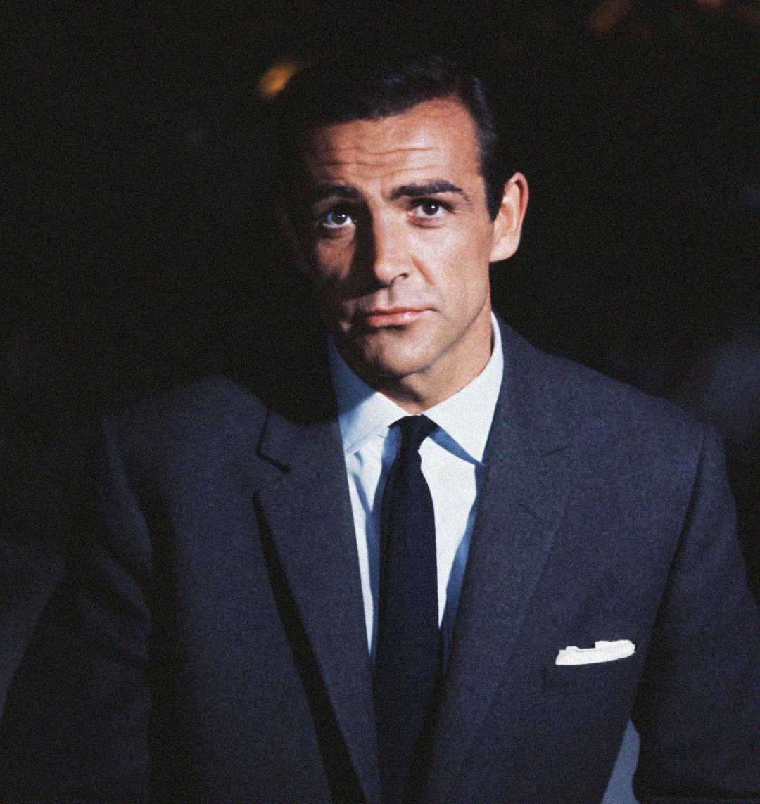 A man with dark hair wearing a dark suit, white shirt, and black tie looks directly at the camera against a dark background. He has a serious expression and a pocket square in his suit jacket.