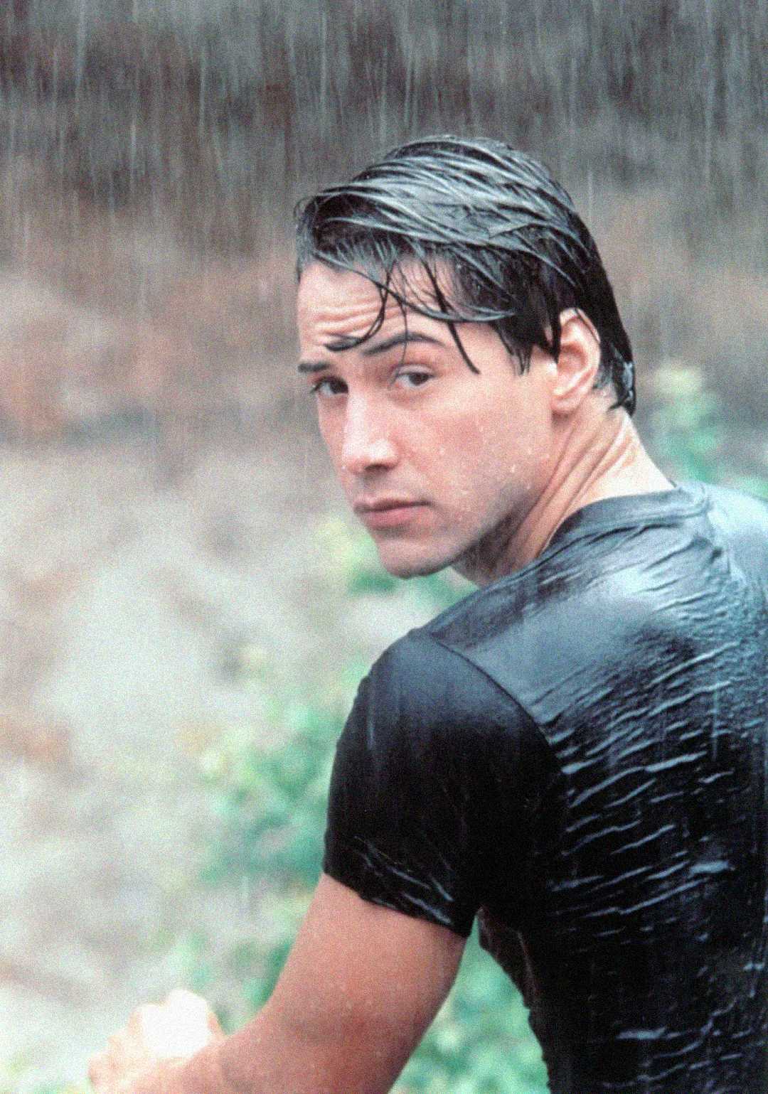A young man with wet dark hair and a soaked black t-shirt looks over his shoulder in the rain, with a blurred natural background behind him.