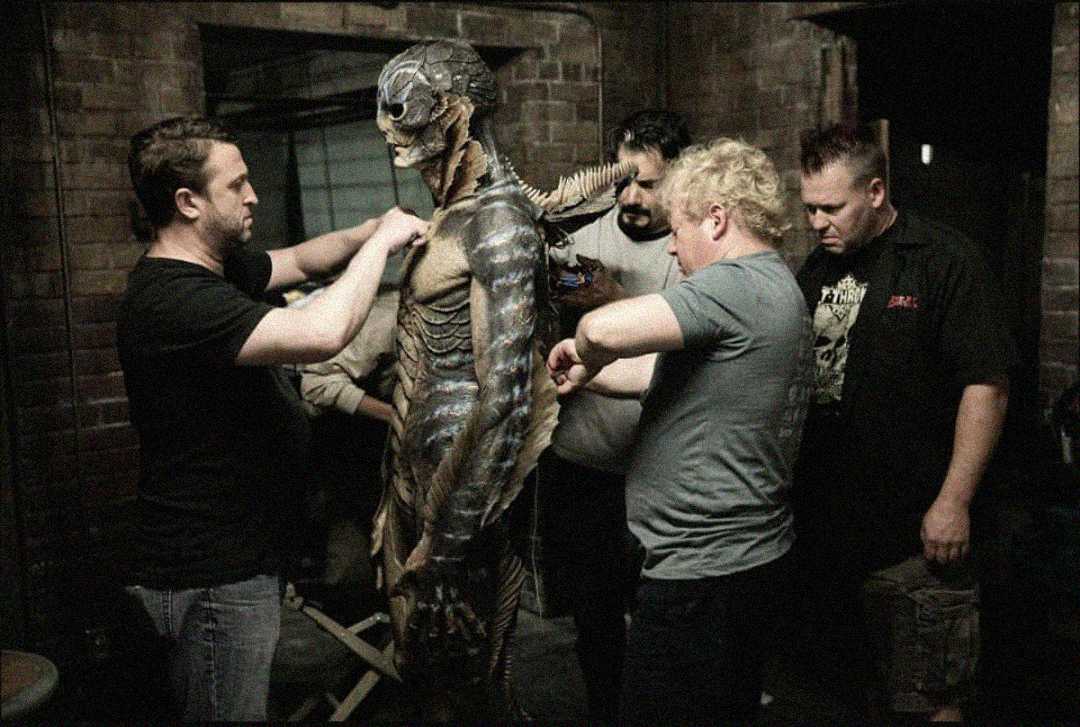 Several people adjust and fit a detailed, alien-like creature costume on an actor in a dimly lit room with brick walls, likely behind the scenes of a movie or television production.