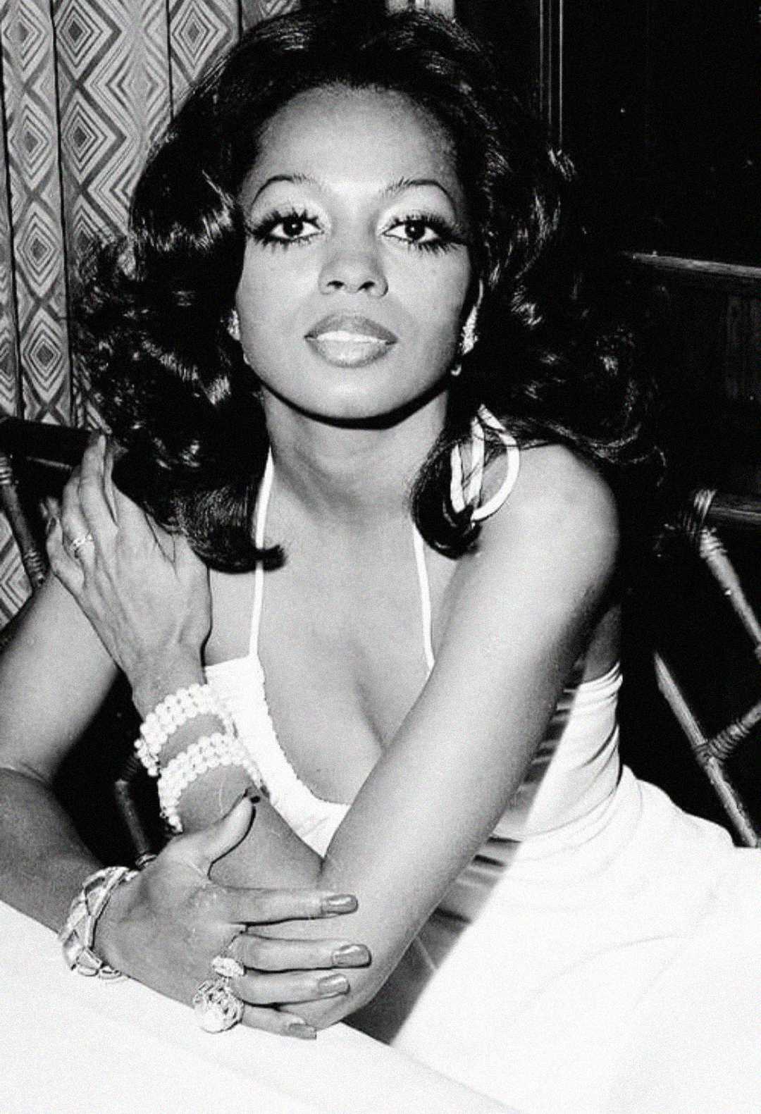 A glamorous woman with voluminous hair poses at a table, wearing a sleeveless dress, hoop earrings, and several bracelets, looking confidently at the camera in this black and white photo.