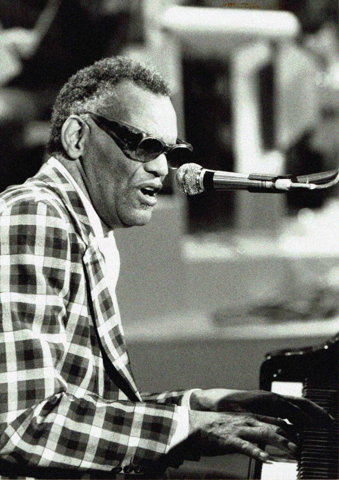A man wearing dark sunglasses and a checkered suit plays the piano and sings into a microphone on stage. The image is in black and white.
