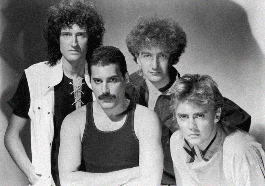 A black-and-white photo of four men with serious expressions, standing close together. Three have curly hair, and one has short hair and a mustache. They are wearing casual 1980s-style clothing.