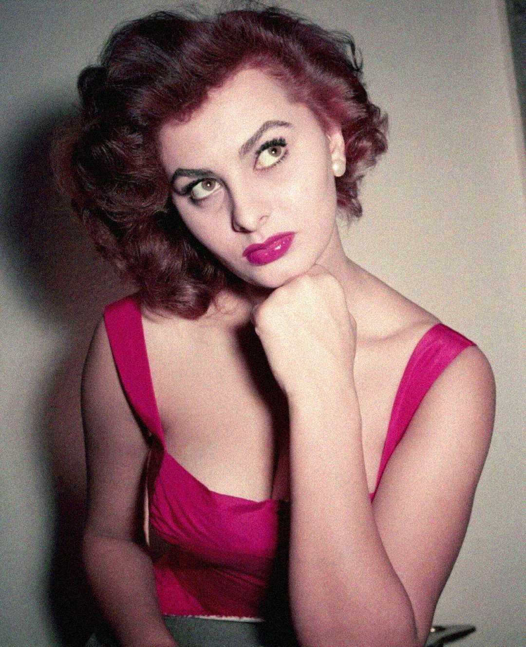 A woman with wavy, dark hair wearing a sleeveless, bright pink top rests her chin on her hand and gazes upward thoughtfully. The background is plain and softly lit.
