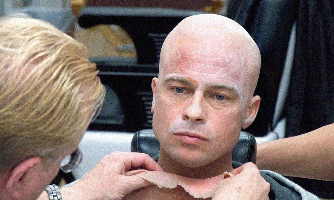 A makeup artist applies a bald cap and prosthetic makeup to a man, transforming his appearance by adding realistic wrinkles and a hairless scalp in a studio setting.