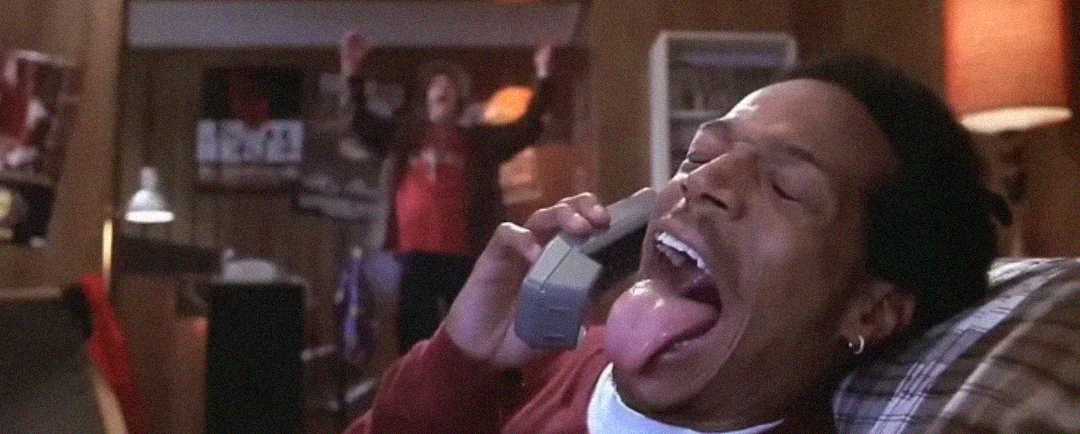 A person reclines on a couch, holding a phone to their ear and sticking out their tongue dramatically. In the background, another person stands with arms raised in a wood-paneled room.