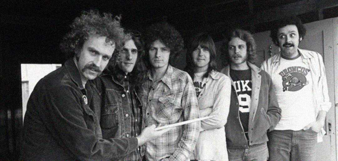 Six men stand in a casual line inside a room, dressed in 1970s-style clothing. The man in front points with a drumstick, while the others stand closely behind, some smiling slightly. The image is in black and white.