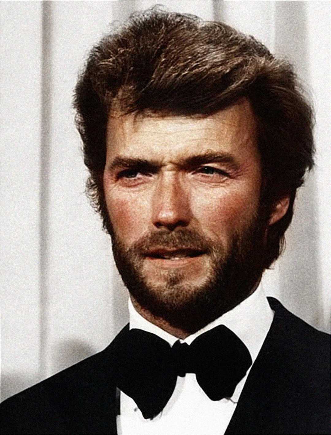 A man with light skin, tousled brown hair, and a thick beard wearing a black tuxedo, bow tie, and white shirt, stands in front of a neutral background.