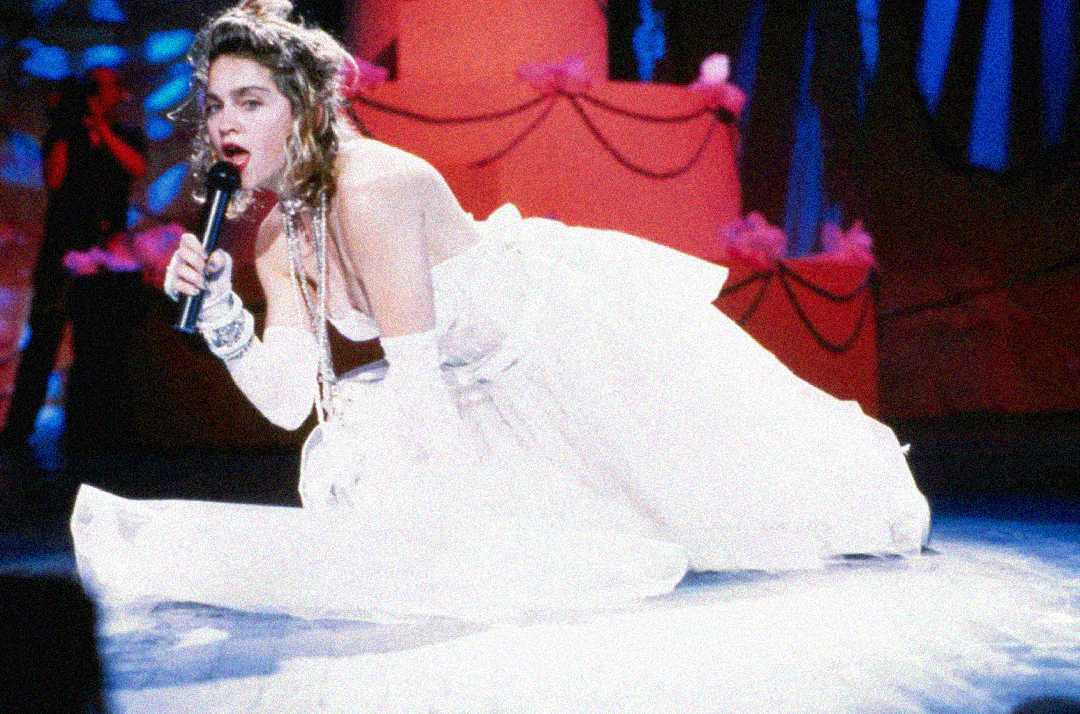 A performer in a white wedding dress and gloves sings passionately into a microphone while kneeling on stage, with red and blue decorations in the background.